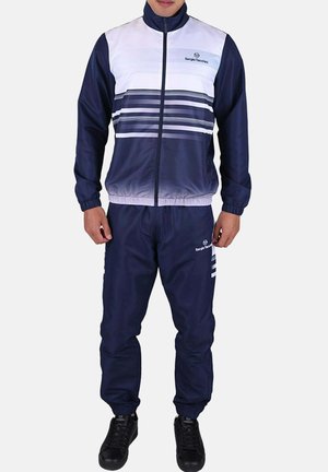 Man wearing a navy and white Sergio Tacchini tracksuit with striped design, standing with hands relaxed by sides against a plain background.