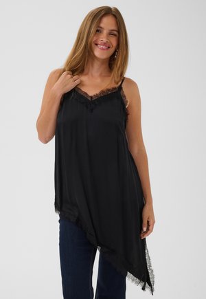 Femme souriante, portant un débardeur en satin noir avec une bordure en dentelle et un ourlet asymétrique, assorti à un jean bleu foncé sur un fond uni.