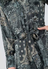 Langmouw zwarte jurk met een paisleypatroon in beige en wit. Voorzien van knopen en een geknoopte taille met geplooide mouwen. Gladde stoftextuur.