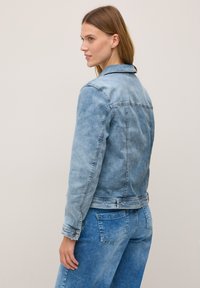 Femme aux longs cheveux portant une veste en denim bleu clair et un pantalon assorti, se tenant devant un fond neutre, tournant légèrement le dos.