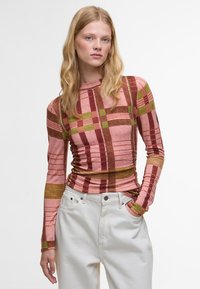 Langärmliges Oberteil mit gerafftem Design in einem karierten Muster aus Rosa, Braun und Grün; kombiniert mit hoch taillierten weißen Hosen.