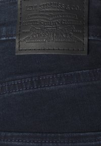 Svart lädermärke med präglad text, fäst vid mörkblå denim, med synliga sömmar och en slät yta.