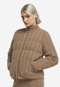 Brun stickad jacka med hög krage, som har ett kabelstickningsmönster, dragkedja och framfickor. Mjuk textur och avslappnad passform.