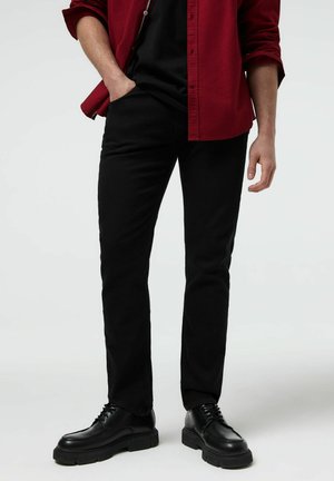 Mężczyzna ubrany w czarne spodnie, czarne buty i czerwoną koszulę na guziki, z jedną ręką w kieszeni na jednolitym tle.