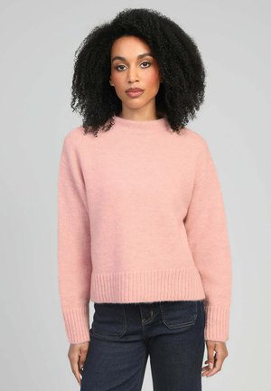 Femme aux cheveux bouclés portant un pull en maille rose clair et un jean bleu foncé, debout devant un fond gris uni.
