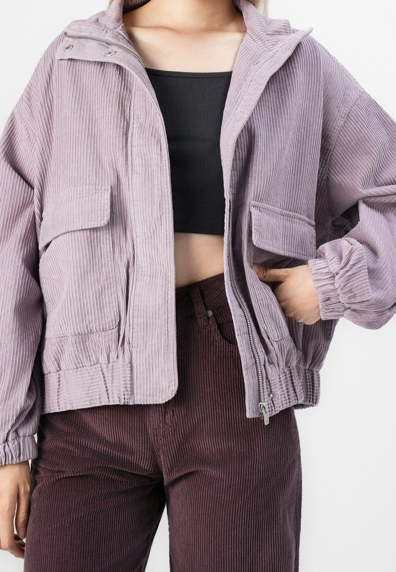 Personne portant une veste en velours côtelé violet clair sur un crop top noir et un pantalon en velours côtelé violet foncé, main reposant sur la poche de la veste.