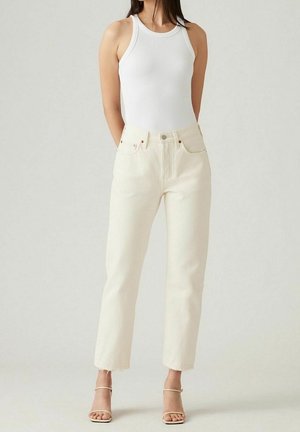 Woman wearing sleeveless white ribbed top, off-white high-waist jeans, and beige strappy heels, standing against a light background.