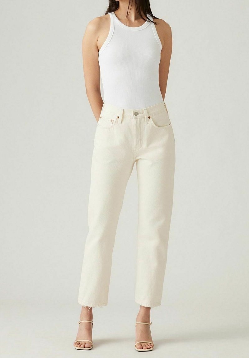 Femme portant un débardeur blanc côtelé, un jean taille haute couleur écru et des talons beiges à lanières, debout devant un fond clair.