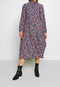 Mörkblå långärmad klänning med ett blommigt paisley-mönster i rosa och vitt, med hög krage och samlad midja, kombinerad med svarta ankelstövlar.