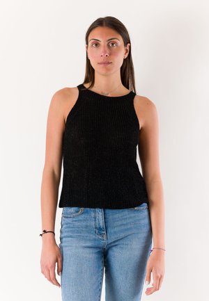 Donna con lunghi capelli castani che indossa una canotta nera e jeans azzurri, in piedi davanti a uno sfondo bianco semplice.