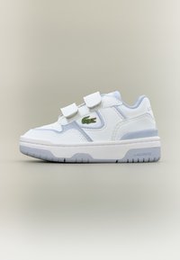 Λευκό παιδικό αθλητικό παπούτσι με δύο ιμάντες βέλκρο, φωτεινή γκρι σόλα και πράσινο λογότυπο κροκόδειλου Lacoste στο πλάι.