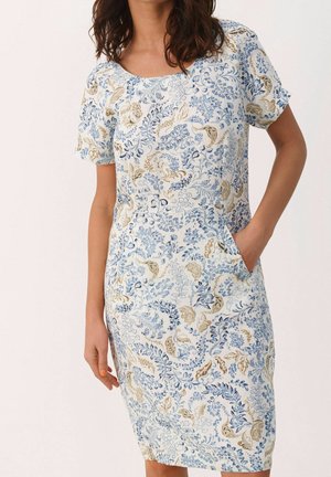 Femme portant une robe blanche mi-longue avec des motifs floraux bleus et beiges et des manches courtes, main dans la poche.