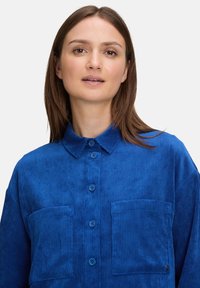 Niebieska koszula z korduroyu z kołnierzykiem, z przodu z zapięciem na guziki oraz z dwiema kieszeniami na piersi. Teksturowany materiał z pionowym prążkowaniem.