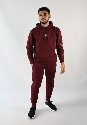 Conjunto de sudadera y pantalones de chándal burdeos, fabricado en algodón, que presenta un bolsillo tipo canguro, detalles con logotipo y puños acanalados, combinado con zapatillas negras.