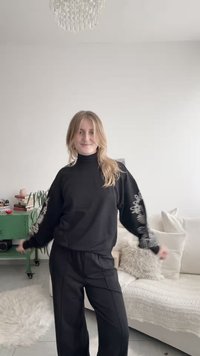 Mladá žena v černém tričku s dlouhým rukávem a kalhotách pózuje v jasném obývacím pokoji s rostlinami, knihami a bílou pohovkou s polštáři.