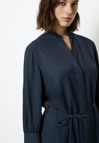 Abito blu navy a maniche lunghe con colletto alla cinese, forma aderente e cintura in vita. Tessuto leggero e liscio con una leggera lucentezza.