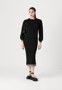 Crna pletena haljina dugih rukava s rebrastim donjim rubom i manžetama. Ima prorez sa strane i kombinira se s debelim crnim cipelama i malom torbom.