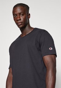 Schwarzes Baumwoll-T-Shirt mit kurzen Ärmeln und Rundhalsausschnitt. Besitzt ein kleines bunt gesticktes Logo auf dem linken Ärmel.