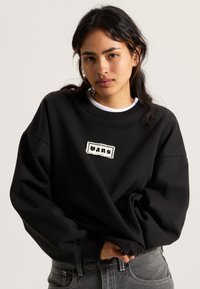 Sweatshirt noir surdimensionné en tissu épais, avec un patch logo brodé blanc centré sur la poitrine. Fini doux et texturé.