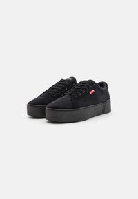 Black suede sneaker with a thick black rubber sole, featuring lace-up design and a small red logo on the side.