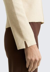 Beżowy sweter dziany z rozcięciem na dole i wyraźną fakturą, w połączeniu z brązowymi dopasowanymi spodniami. Zbliżenie na detal rękawa i dłoni.