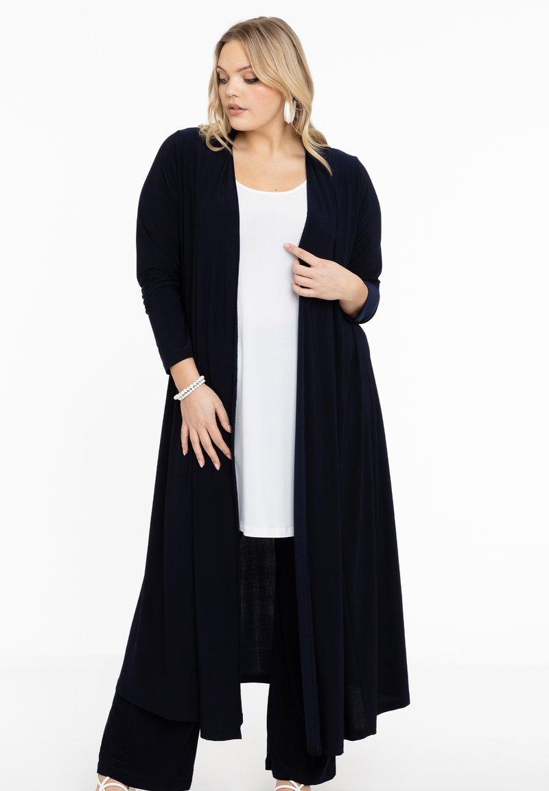 Langer Marineblauer Cardigan aus weichem Stoff, mit offenem Frontdesign, kombiniert mit einem weißen Oberteil und dunklen Hosen, mit glatter Textur und fließender Silhouette.