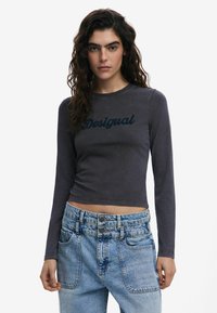 Dlhý rukáv šedý top s tmavomodrým logom "Desigual". Skrátený strih, hladká textúra. Nosí sa s svetlomodrými riflami s vysokým pásom.