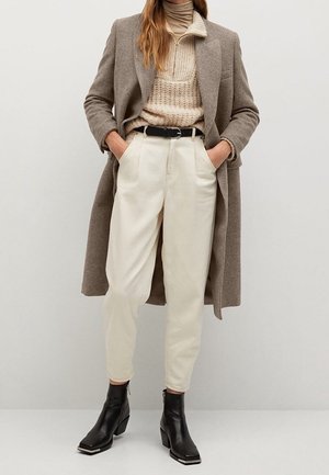 Kvinde iført beige rullekravesweater, cremehøjtaljede bukser, brun uldfrakke, sort bælte og sorte ankelstøvler, stående med hænderne i lommerne.