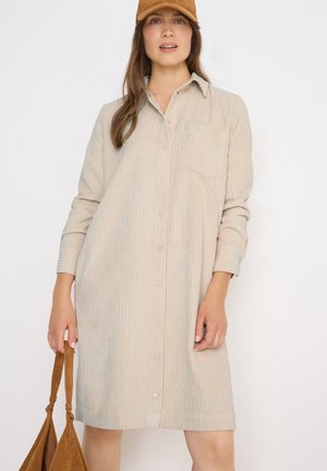 Robe chemise en velours côtelé beige clair avec manches longues, devant à boutons, poche poitrine et coupe décontractée. Portée avec une casquette marron et un sac à main.