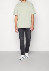 Persona con una camiseta de gran tamaño de color verde claro, vaqueros negros ajustados y zapatillas blancas con detalles verdes y naranjas, de pie sobre un suelo gris.