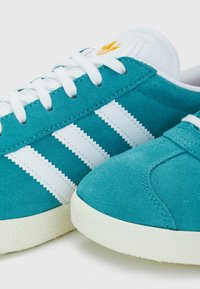 Baskets en daim turquoise avec trois bandes blanches, bout rond et semelle en caoutchouc texturée couleur blanc cassé. Caractéristiques : lacets blancs et languette avec logo.