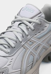 Iš arti balta ir šviesiai pilka Asics sportbačio su išsamia raištelių, tinklelio audinio ir firminio logotipo šone bei liežuvėlyje nuotrauka.