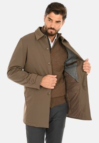 Cappotto marrone di lunghezza media con colletto e chiusura con bottoni, con interno foderato in grigio e tasca sul petto. Tessuto liscio, vestibilità sartoriale.