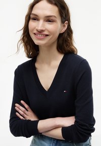 Smilende kvinde med bølget brunt hår iført en sort V-hals sweater med lille logo og blå jeans, armene krydset.