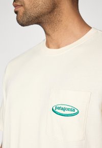 Μπλούζα από βαμβάκι σε κρεμ απόχρωση με τσέπη στο στήθος που φέρει κεντημένο μπλε-πράσινο λογότυπ ο «patagonia». Κλασικός στρογγυλός γιακάς και κοντά μανίκια.