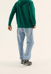 Sudadera con capucha verde, jeans de mezclilla azul claro y zapatillas deportivas negras con acentos grises, vistas desde la espalda contra un fondo neutro.