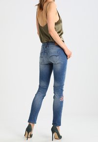 Slim-fit blå denimjeans med lätt slitage och blommig broderi på höger lår; matchade med en slät olivgrön topp och högklackade skor.