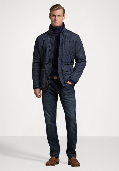 Giacca trapuntata blu navy con colletto alto, bottoni frontali e tasche laterali, abbinata a un maglione scuro, jeans in denim e scarpe di pelle marroni.