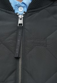 Chaqueta bomber negra acolchada con cremallera plateada, con cuello acanalado y un sutil patrón de costuras horizontales. Logo en el tirador de la cremallera.