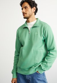 Homem a usar um moletom verde de zip com um camisola branca por baixo, com as mãos nos bolsos de uns jeans azuis, olhando para o lado contra um fundo branco.