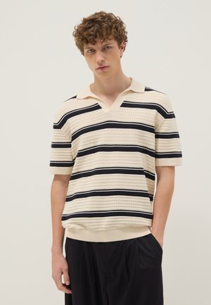 Joven con pelo rizado que lleva una camisa polo de punto color crema con rayas horizontales negras y pantalones negros holgados.