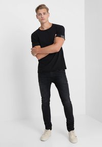 Mustat lyhyet hihattomat t-paidat, joissa on teksturoitu olkatieto, yhdistetty tummiin slim-fit-farkkuihin ja vaaleisiin lenkkareihin yksinkertaisella taustalla.