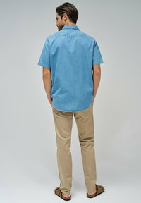 Camisa de denim de manga corta azul claro con dobladillo redondeado, combinada con pantalones beige y sandalias marrones, vista desde la parte de atrás.