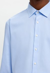 Ανοιχτό μπλε πουκάμισο με μακριά μανίκια και γιακά με κουμπιά, με λευκά κουμπιά και λεία, υφαντή υφή.
