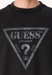 Melna džemperis ar apaļu izgriezumu, ar lielu pelēku trīsstūra logo, kurā ir teksts "GUESS ORIGINALS" un jautājuma zīmes dizains.