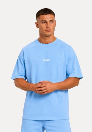 Mand med kort hår iført en lyseblå oversize t-shirt og matchende shorts, står med hænderne foldet og kigger til siden.