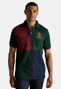 Polo krekls ar krāsu bloku dizainu zaļā, tumši sarkanā un tumši zilā krāsā. Iekļauj izšūtus zīmolus, divu pogu aizdari un teksturētu apdari.