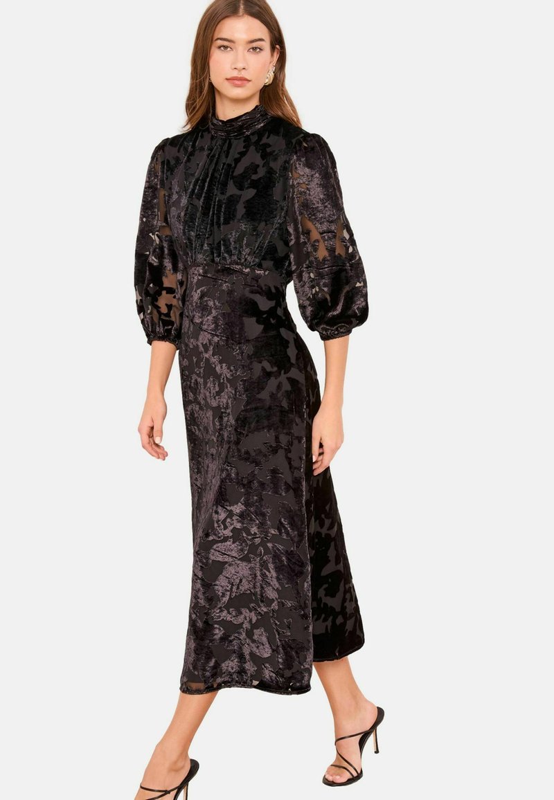 Robe noire à motif floral avec manches bouffantes transparentes et col montant. Texture lisse, longueur jusqu'aux chevilles, silhouette ajustée, portée avec des talons à brides.