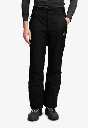 Personne portant un pantalon de plein air noir avec une poche cargo latérale et des chaussures de randonnée noires, debout avec une main dans la poche.