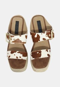 Sandalias planas con estampado de vaca, con tira de ante marrón y fondo blanco, que presentan una hebilla dorada y una suela intermedia de yute.